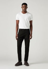 Tânărul stă în picioare, purtând un tricou alb cu mânecă scurtă, pantaloni negri slim-fit și pantofi împletiți de culoare maro închis, pe un fundal deschis uniform.