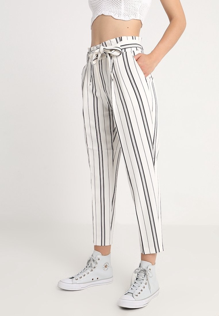 Černobílé pruhované zkrácené kalhoty s páskem v pase, vyrobené z lehké tkaniny, v kombinaci s vysokými teniskami.