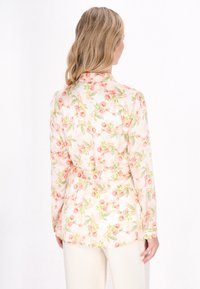 Blomstret bluse i lys fersken med lange ærmer, der har et blomstermønster af lyserøde roser og grønne blade, lavet af et gennemsigtigt og blødt stof.