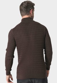 Brązowy sweter z dzianiny z wyraźnym wzorem w warkocze, ściągaczami przy mankietach i wysokim kołnierzem. Materiał wydaje się miękki i gęsty.