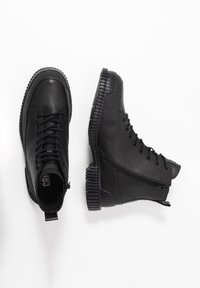 Par de botas de cuero negro hasta el tobillo con suelas gruesas, cordones en la parte delantera, cremalleras laterales y lengüetas de agarre sobre un fondo blanco.