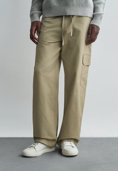 Pantaloni cargo beige con una vestibilità rilassata, realizzati in tessuto leggero, dotati di tasche laterali e cintura con cordino, abbinati a sneakers bianche.