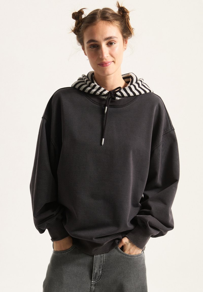 Overmaat zwart sweatshirt met een gestreepte grijs-witte capuchon. Beschikt over een trekkoord en een textuurstof, met brede mouwen en een ontspannen pasvorm.