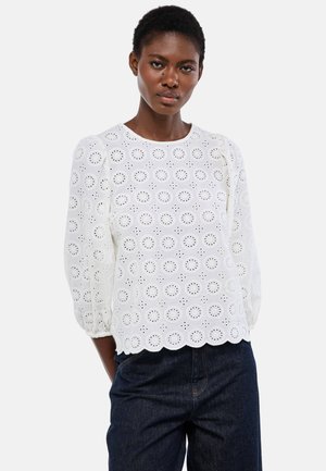 Femme portant un chemisier en dentelle œillet blanche avec des manches 3/4 bouffantes et un ourlet festonné, associé à un jean bleu foncé.