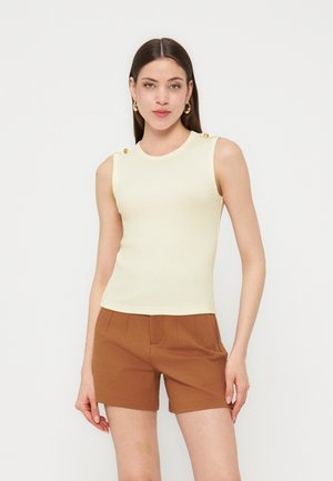 Donna con capelli castani lisci che indossa un top crema senza maniche con bottoni dorati e pantaloncini marroni, in piedi davanti a uno sfondo bianco.