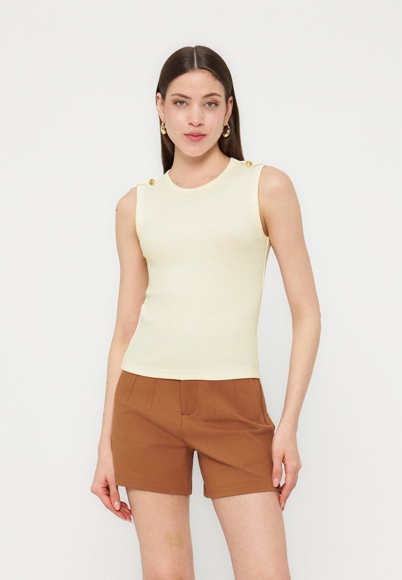 Γυναίκα με ίσια καστανά μαλλιά, που φοράει αμάνικο κρεμ τοπ με χρυσά κουμπιά και καφέ σορτς, στέκεται μπροστά σε λευκό φόντο.