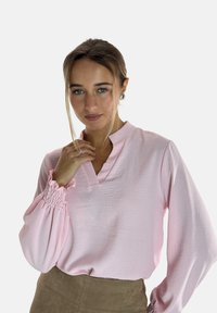 Lichtroze blouse met lange mouwen, voorzien van een geplooide kraag en elastische manchetten. Getextureerde stof en een relaxed fit. Gestyleerd met een bruine rok.