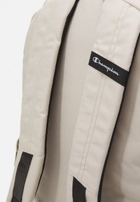 Beige canvas backpack with black accents, featuring padded shoulder straps and a Champion logo label on one strap. Textured surface.