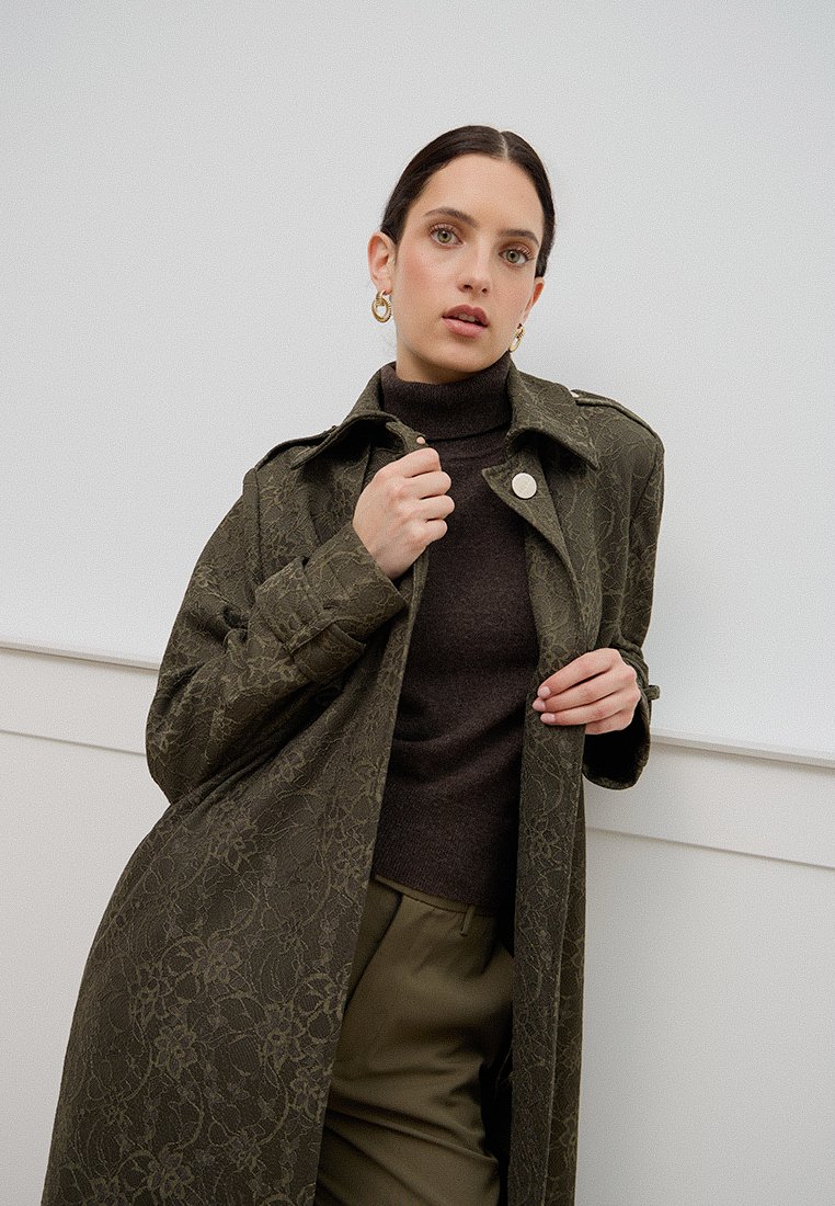 Cappotto lungo verde oliva con motivo in pizzo, caratterizzato da un colletto a revers e chiusura a bottone, indossato sopra un dolcevita marrone scuro e pantaloni verdi.