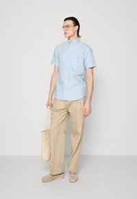 Camisa azul clara de manga curta com colarinho abotoado, calças bege e uma bolsa tote bege clara. Usada com sapatos cinza claro.