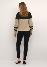 Pull en tricot beige et noir avec des accents rayés aux épaules et aux poignets. Associé à un pantalon noir ajusté et des chaussures à talons hauts.
