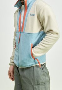 Giacca in pile con parte superiore beige e parte inferiore azzurra chiara, accentuata da cerniere e tasche arancioni, colletto con zipper e texture morbida.