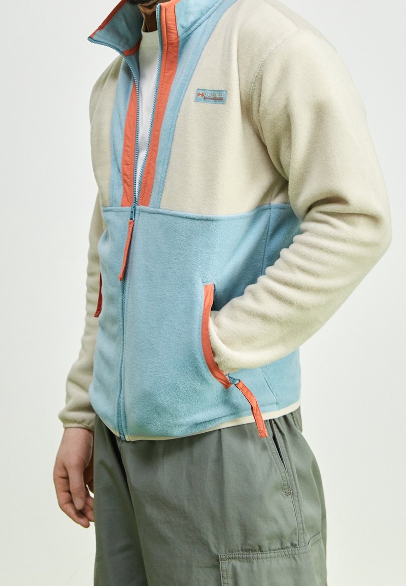 Giacca in pile con parte superiore beige e parte inferiore azzurra chiara, accentuata da cerniere e tasche arancioni, colletto con zipper e texture morbida.