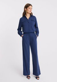 Kobieta w granatowej koszuli zapinanej na guziki i szerokich spodniach z rękami w kieszeniach, stojąca na tle jednolitego białego tła.