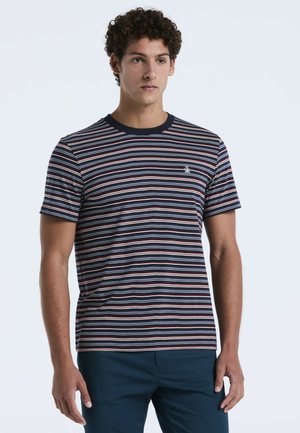 Junger Mann steht mit einem kurzärmeligen T-Shirt in Marineblau, Weiß und Rot mit gestreiften Muster und dunkelbblauen Hosen vor einem schlichten hellen Hintergrund.