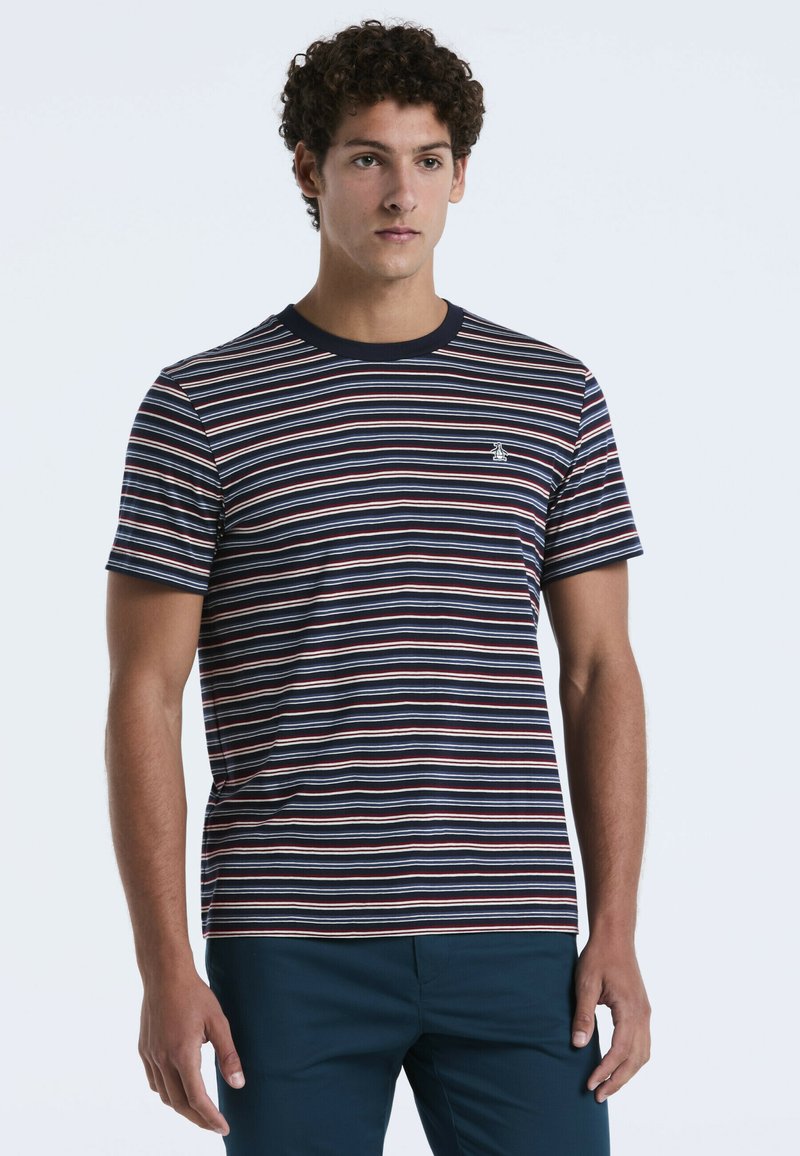 Jonge man staat in een marineblauwe, witte en rode gestreepte t-shirt met korte mouwen en donkere blauwe broek tegen een effen lichte achtergrond.