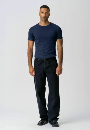 Mand stående vendt fremad iført en mørkeblå t-shirt, sorte bukser med vide ben og sorte sko mod en ensartet lys baggrund.