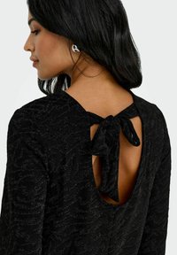 Svart teksturert topp med lav rygg og detalj med knytt sløyfe. Stoffet har et subtilt mønster og en behagelig, avslappet passform.