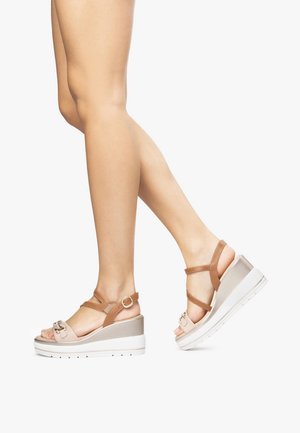 Jambes féminines portant des sandales compensées beige et marron avec des brides à la cheville, debout sur un fond blanc.