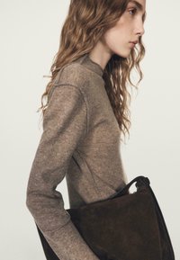 Junge Frau mit welligem Haar, die einen braunen Strickpullover trägt und eine große dunkelbraune Wildleder-Handtasche unter dem Arm hält.