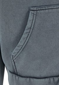 Gros plan d'une poche en tissu gris avec couture visible et ceinture froncée, probablement partie d'un sweat-shirt ou d'un pantalon de survêtement.