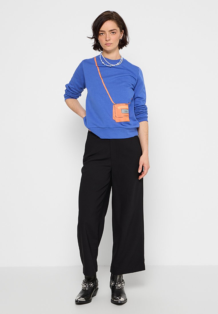 Sudadera azul de mangas largas, diseño de bolso cruzado naranja, combinada con pantalones anchos negros y botines brillantes negros.
