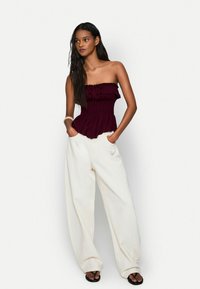 Top bordeaux senza spalline con bordi arricciati e busto aderente, abbinato a pantaloni bianchi larghi e infradito nere. Texture del tessuto liscia.