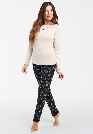 Beżowa koszula z długim rękawem, okrągłym dekoltem i czarnym akcentem. Czarne spodnie z wzorem w złote i białe gwiazdy. Miękka tkanina. Modelka stoi boso.