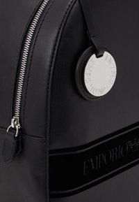 Black leather backpack featuring a silver zipper and a round metallic tag embossed with "Emporio Armani." Textured surface with sleek design.