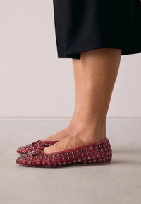 Ballerines bordeaux avec des œillets métalliques et un nœud à l'avant. Matière douce, forme arrondie et profil bas, adaptées à un port décontracté.