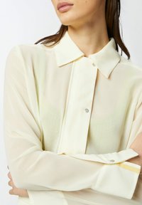 Camicia gialla trasparente con colletto classico, maniche lunghe e chiusura con bottoni. Presenta dettagli con bottoni argentati e polsini a righe gialle.