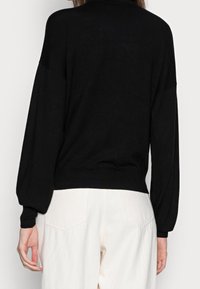 Pull en maille noire avec un ourlet ajusté et des manches bouffantes. Texture douce. Porté avec un pantalon clair doté de poches arrière.