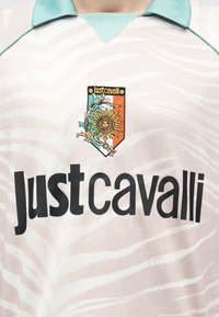 Ανοιχτό πουκάμισο με ρίγες ζέβρας, γιακά σε τιρκουάζ απόχρωση, μεγάλο μαύρο κείμενο "Just Cavalli" και μικρό κεντητό έμβλημα με σχέδιο ήλιου και λουλουδιών.