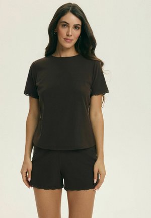 Kobieta o długich brązowych włosach ubrana jest w prostą ciemnobrązową koszulkę z krótkim rękawem oraz dopasowane spodenki na neutralnym tle.