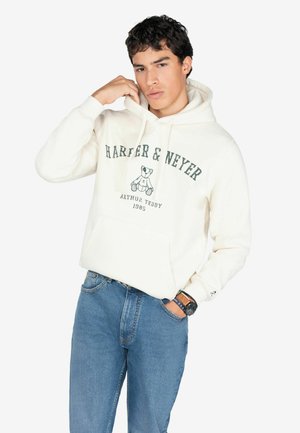 Sudadera color crema con un gráfico de un oso de peluche y el texto "HARPER & NEYER" en verde oscuro. Combinada con jeans de mezclilla azul claro.