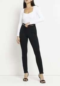 Witte bodysuit met lange mouwen en een vierkante halslijn, gecombineerd met strakke zwarte broek met een gouden cirkelriem en slanke zwarte hakken.