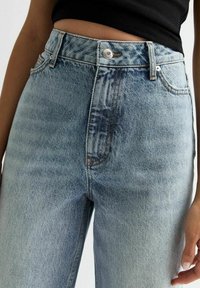 Jeans de mezclilla azul claro de talle alto con un acabado ligeramente desgastado, que cuentan con un cierre de botón, trabillas para cinturón y un diseño tradicional de cinco bolsillos.