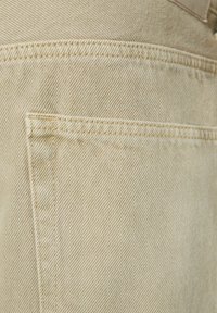 Tessuto in denim beige con trama a costina diagonale. Presenta una singola tasca a toppa e cuciture visibili lungo i bordi.