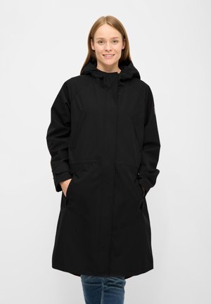 Jonge vrouw die een lange zwarte mantel met kap draagt, handen in de zakken, glimlachend, staand voor een egale lichte achtergrond.