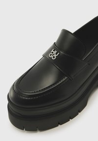 Black leather loafers with a thick sole, featuring a strap with the word "HUGO" in silver. Smooth texture and rounded toe design.