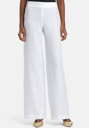 Vrouw draagt witte linnen broek met wijde pijpen en gouden pumps met spits toelopende neus, staande tegen een lichte achtergrond.