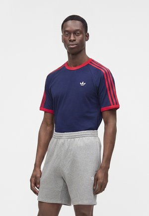 Mladý muž v tmavomodrom tričku Adidas s červenými pásmi a sivých šortkách Adidas stojí pred jednoliatym bielym pozadím.