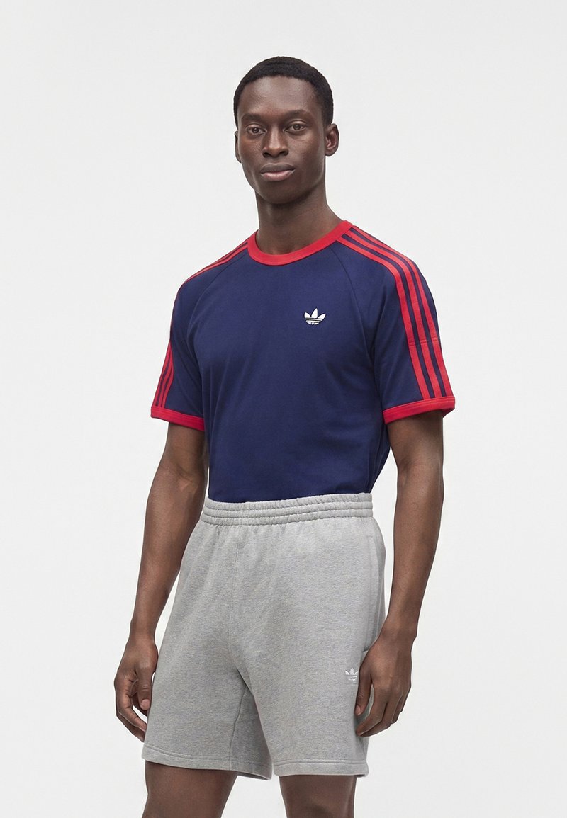Νέος άνδρας φορώντας μπλε σκούρο μπλουζάκι Adidas με κόκκινες ρίγες και γκρι σορτσάκια Adidas στέκεται μπροστά σε λευκό φόντο.