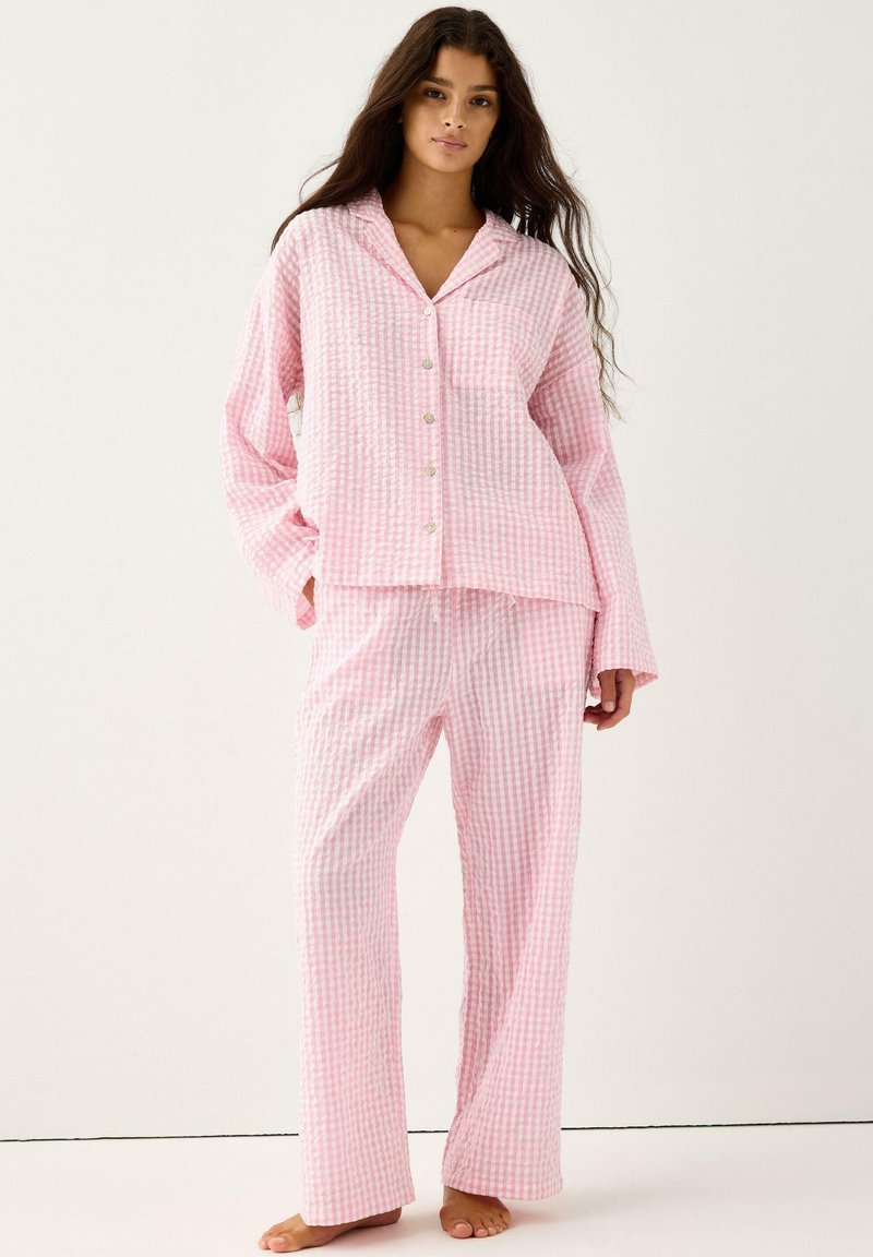 Donna con capelli lunghi che indossa un pigiama a quadri rosa e bianco, largo e con bottoni, in piedi a piedi nudi contro uno sfondo neutro.