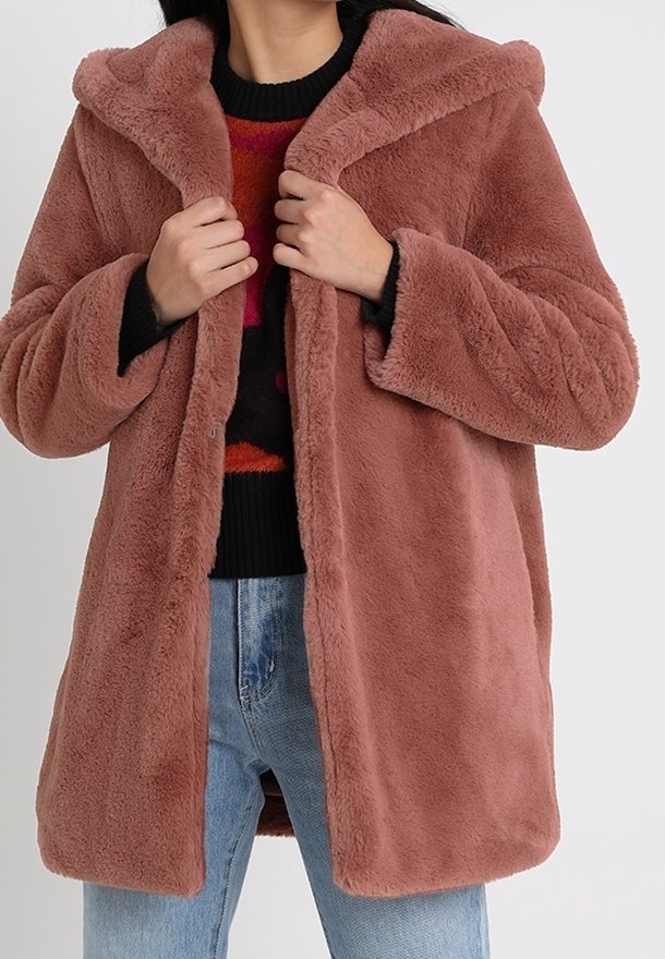 Personne portant un manteau en fausse fourrure brun rose par-dessus un pull multicolore et un jean bleu, tenant les revers du manteau.