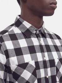 Gros plan d'une personne portant une chemise à carreaux noire et blanche boutonnée, avec col et poches poitrine, tournée vers la droite sur un fond uni.