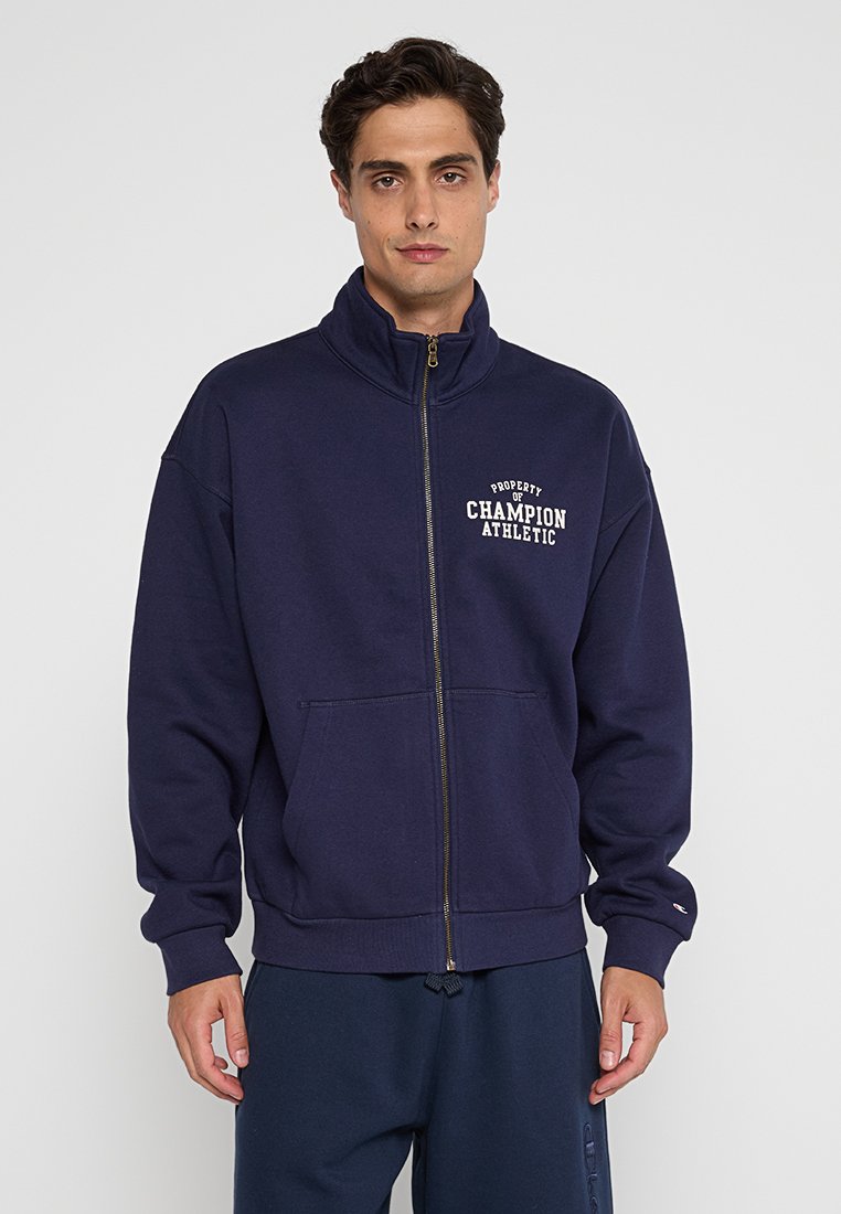 Felpa con zip di colore blu navy realizzata in materiale felpato, con colletto alto, tasche frontali e testo bianco "PROPRIETÀ DI CHAMPION ATHLETIC."