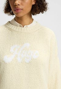Maglione lavorato a maglia color crema con vestibilità ampia, caratterizzato da un logo bianco in un font giocoso al centro e un colletto a costine.