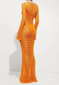 Femme portant une longue robe en tricot ajouré orange montrant la peau en dessous, se tenant contre un fond blanc uni, tournée le dos.