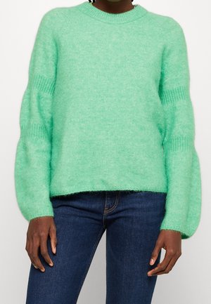 Personne portant un pull duveteux vert clair avec des manches ballon et un jean bleu foncé, debout devant un fond uni.
